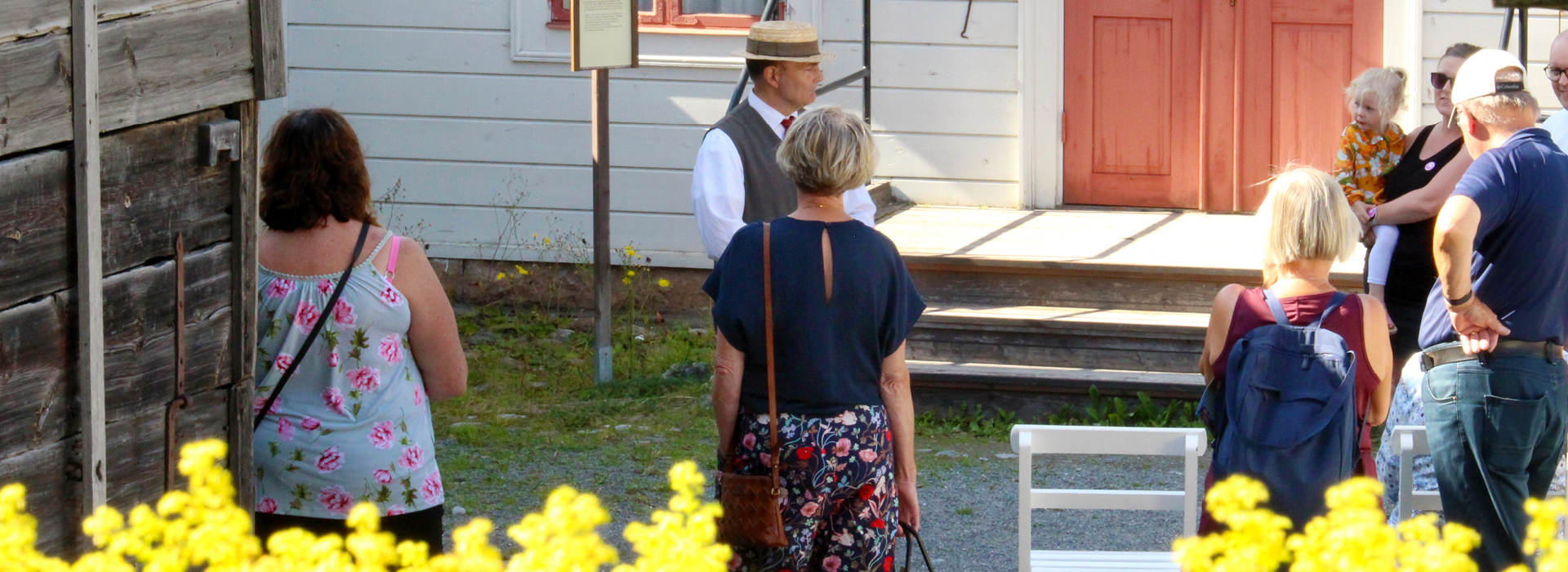 En guide i tidstypiska kläder berättar för en grupp besökare framför en äldre byggnad med röda dörrar och fönster. I förgrunden syns gula blommor.
